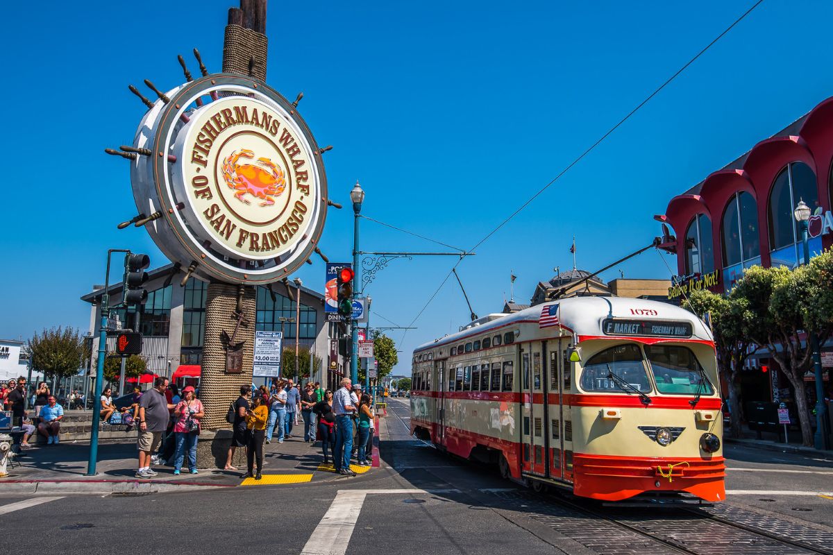 A vintage cream and red F Market streetcar headed to Fisherman's Wharf passes the iconic ship wheel landmark sign on a sunny day, with crowds of tourists gathered on the sidewalk and the Ripley's Believe It or Not building visible on the right.