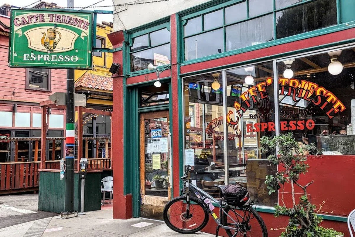 Street-level view of Caffe Trieste, a historic espresso bar in San Francisco's North Beach neighborhood, featuring a green and gold hanging sign, red and green facade with large windows painted with the cafe name, an open door, and a bicycle parked out front.