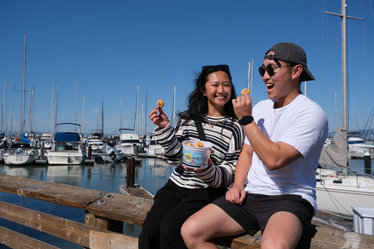 A smiling couple sits on a wooden dock railing eating clam chowder bites from a San Francisco branded cup, with a marina full of sailboats and a clear blue sky stretching behind them at Fisherman's Wharf.