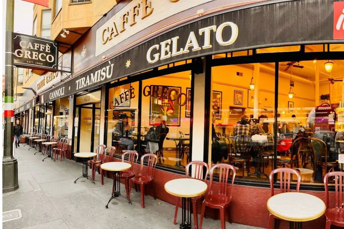 Exterior of Caffe Greco in San Francisco's North Beach, with a curved black storefront awning advertising gelato, tiramisu, and cannoli, rows of empty round tables with red metal chairs lining the sidewalk, and a warmly lit interior visible through large windows.