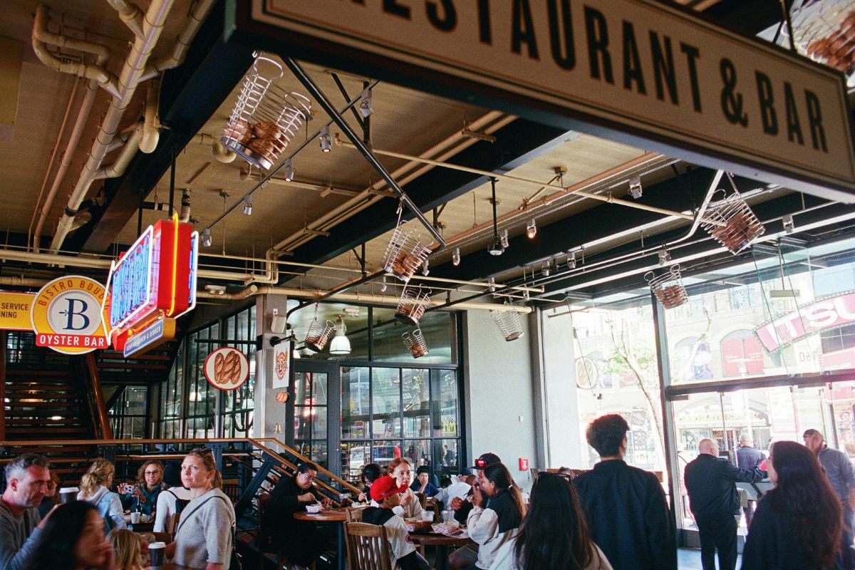 A lively indoor dining scene at a Fisherman's Wharf restaurant and bar, with wire basket light fixtures hanging from an industrial ceiling, neon signs for Bistro Boudin and an oyster bar, and groups of diners at wooden tables beside large windows overlooking the street.
