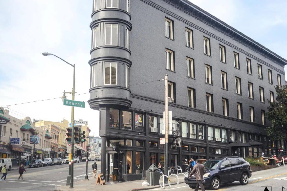 A bold dark gray multi-story historic brick building with a distinctive rounded corner tower sits at the intersection of Kearny Street in San Francisco, with street signs, traffic lights, pedestrians, and a mix of colorful older buildings visible along the busy street behind it.