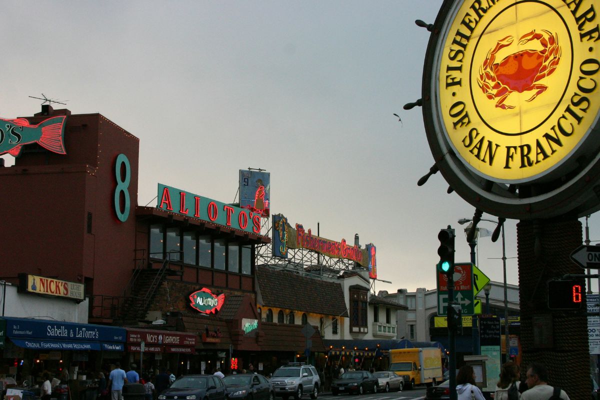 The illuminated Fisherman's Wharf of San Francisco ship wheel sign glows in the foreground at dusk, with the neon-lit facades of Alioto's, Fisherman's Grotto, Nick's Lighthouse, and Sabella and LaTorre's restaurants lining the busy street behind it under a gray evening sky.