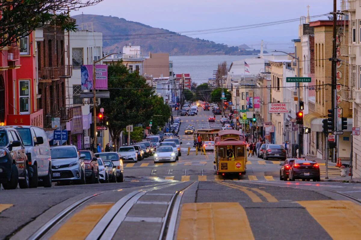 A cable car travels down a steep San Francisco street toward the San Francisco Bay and a hilly island in the distance, flanked by Chinatown storefronts, colorful mixed-use buildings, traffic, and glowing red traffic lights under a dusky purple sky.