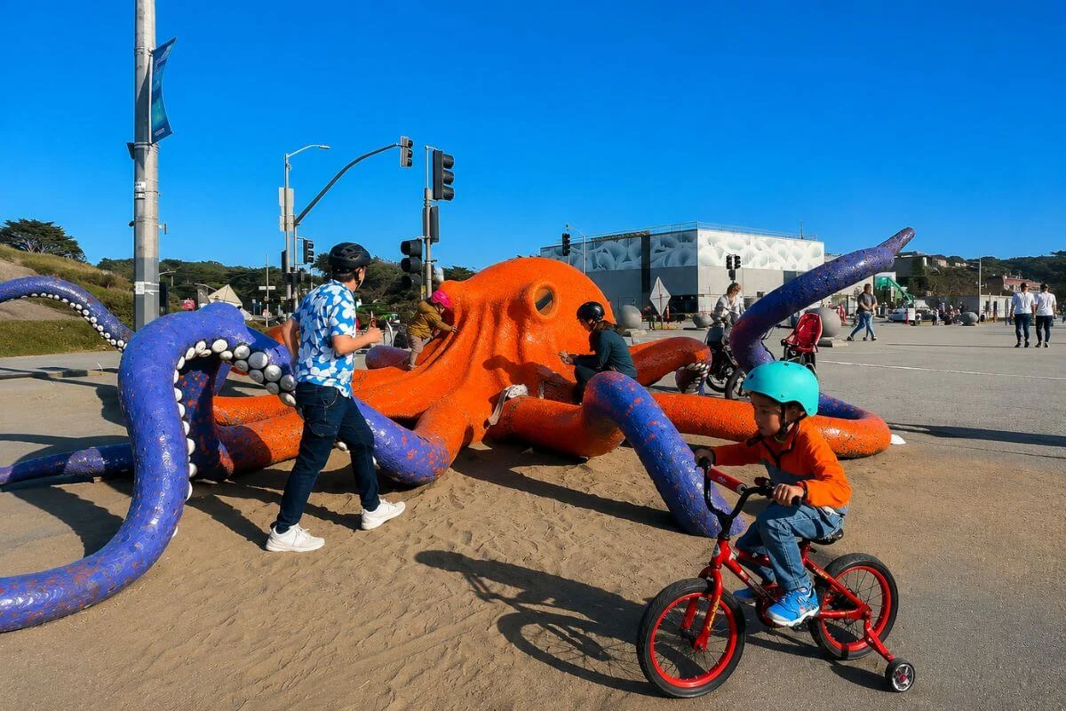 Children and families play around a large orange and purple octopus public art sculpture in a sandy play area near Ocean Beach, San Francisco, with a young boy riding a red bike with training wheels in the foreground.