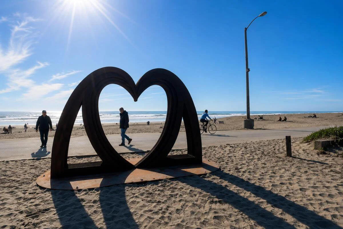 Large metal heart sculpture on sandy beach with walkers and ocean waves in background
