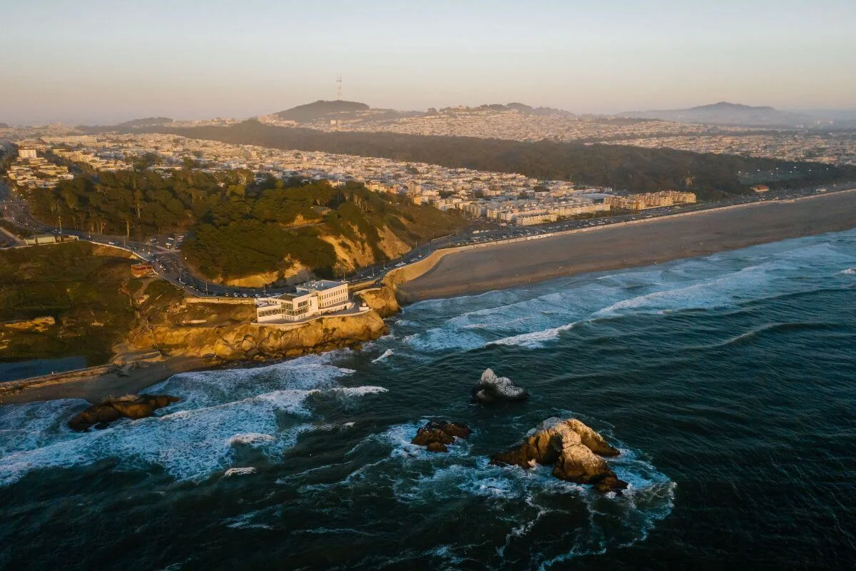 A drone shot of Ocean Beach in San Francisco at golden hour, showing the long stretch of dark sand coastline, crashing waves, rocky outcrops, the Cliff House perched on the headland, and the dense residential neighborhoods of the Outer Sunset stretching inland.