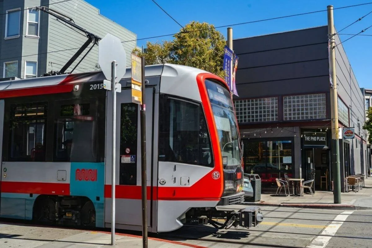 A modern red and white SF Muni light rail train travels along a street in the Outer Sunset neighborhood, with The Pho Place restaurant and low-rise commercial buildings visible in the background under a bright blue sky.