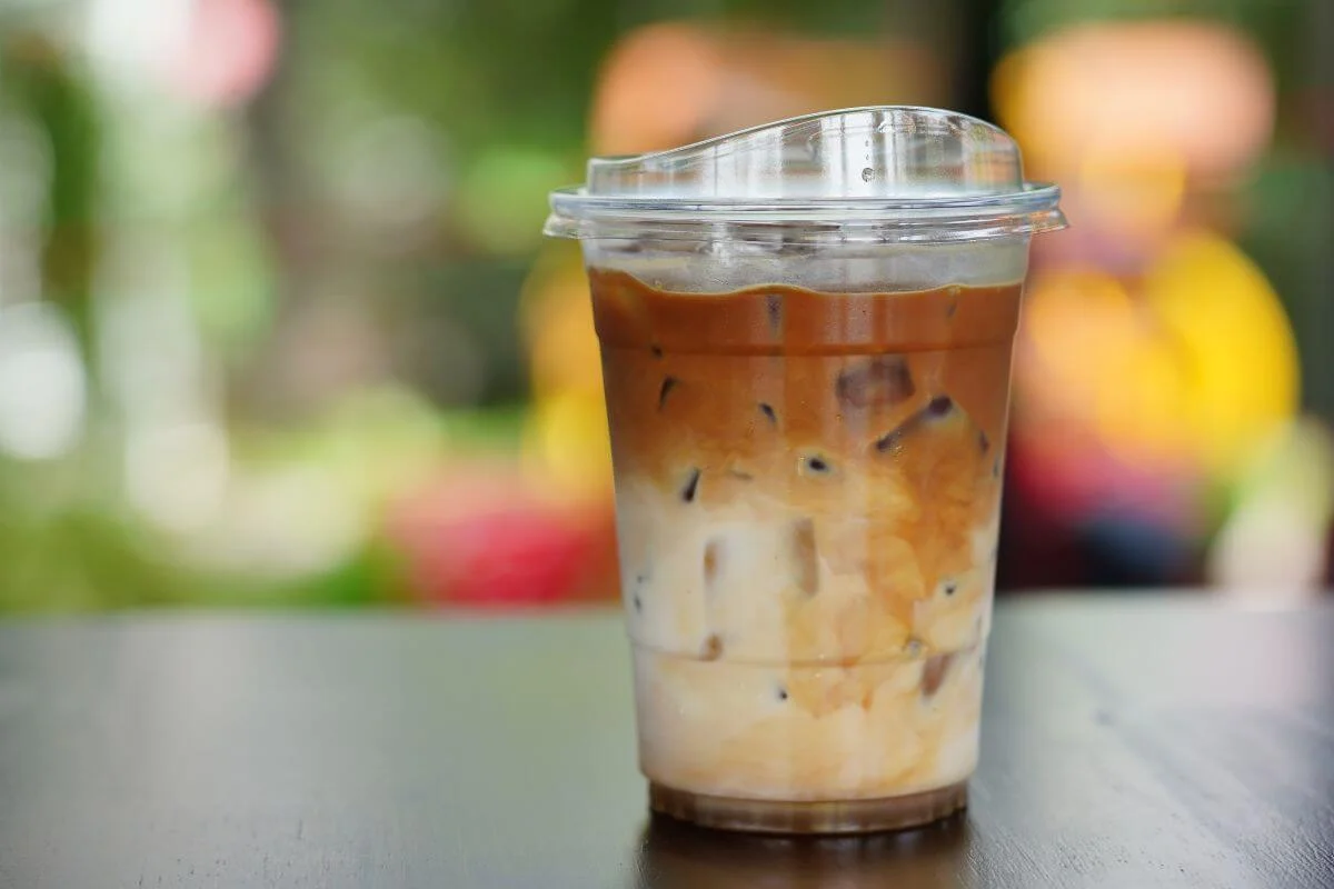 A plastic takeaway cup of iced latte with layered milk and coffee over ice sits on a dark table with a blurred colorful outdoor background.