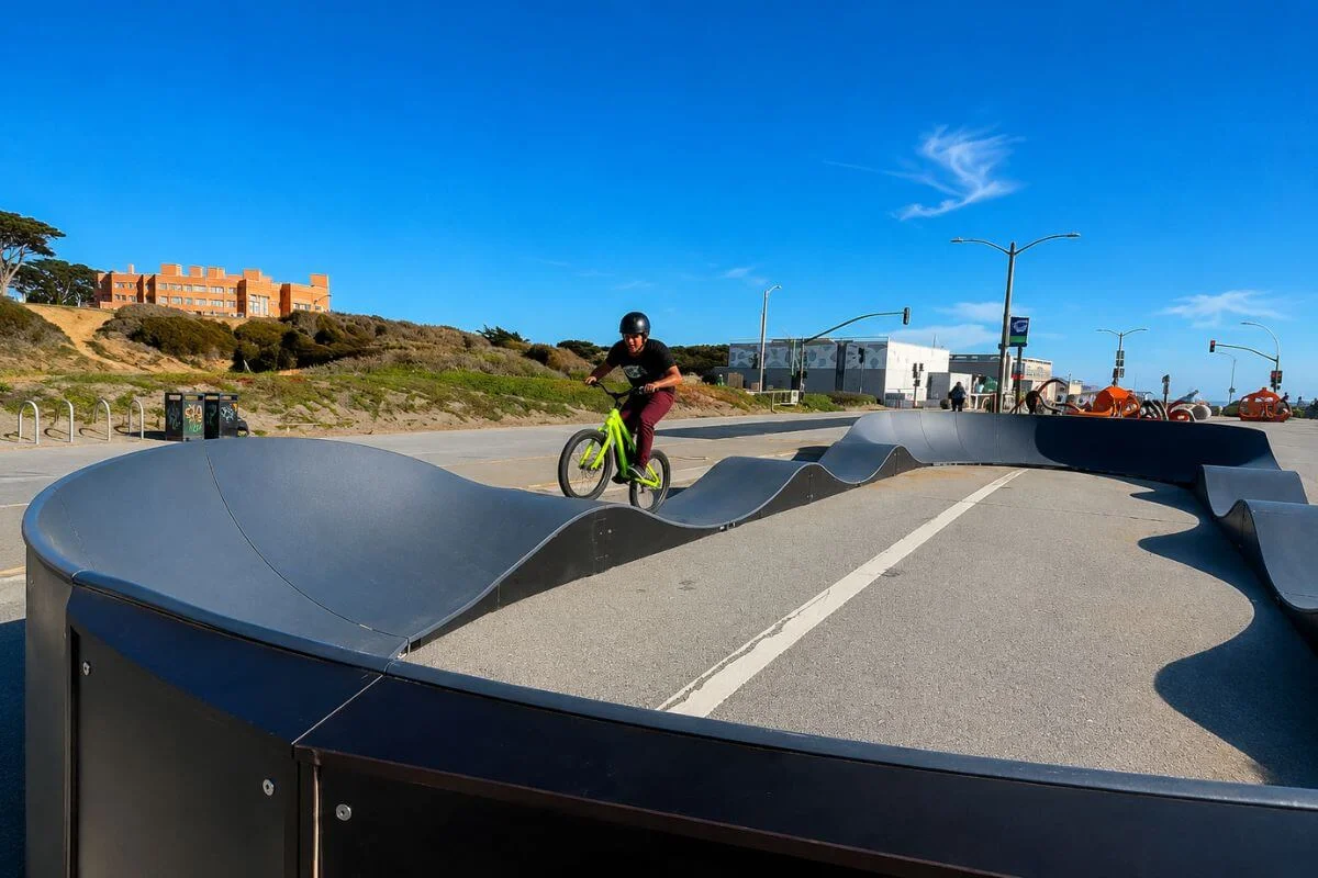 A helmeted BMX rider on a green bike navigates a black pump track near Ocean Beach, with sand dunes, a brick building on the hillside, and the giant octopus sculpture visible in the background under a bright blue sky.