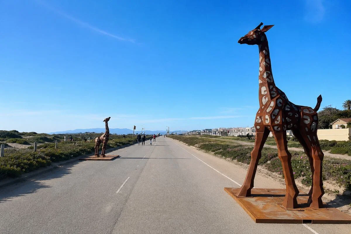Two large rusted steel giraffe sculptures stand on either side of a closed stretch of the Great Highway at Ocean Beach, with pedestrians walking the car-free road and coastal vegetation and residential buildings visible in the distance under a clear blue sky.
