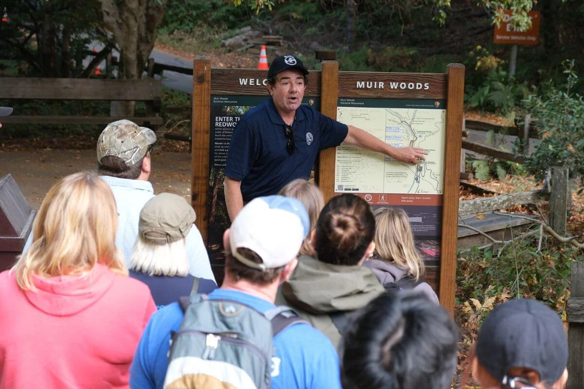 Tour guide pointing at the Muir Woods trail map board while guests gather around at the entrance