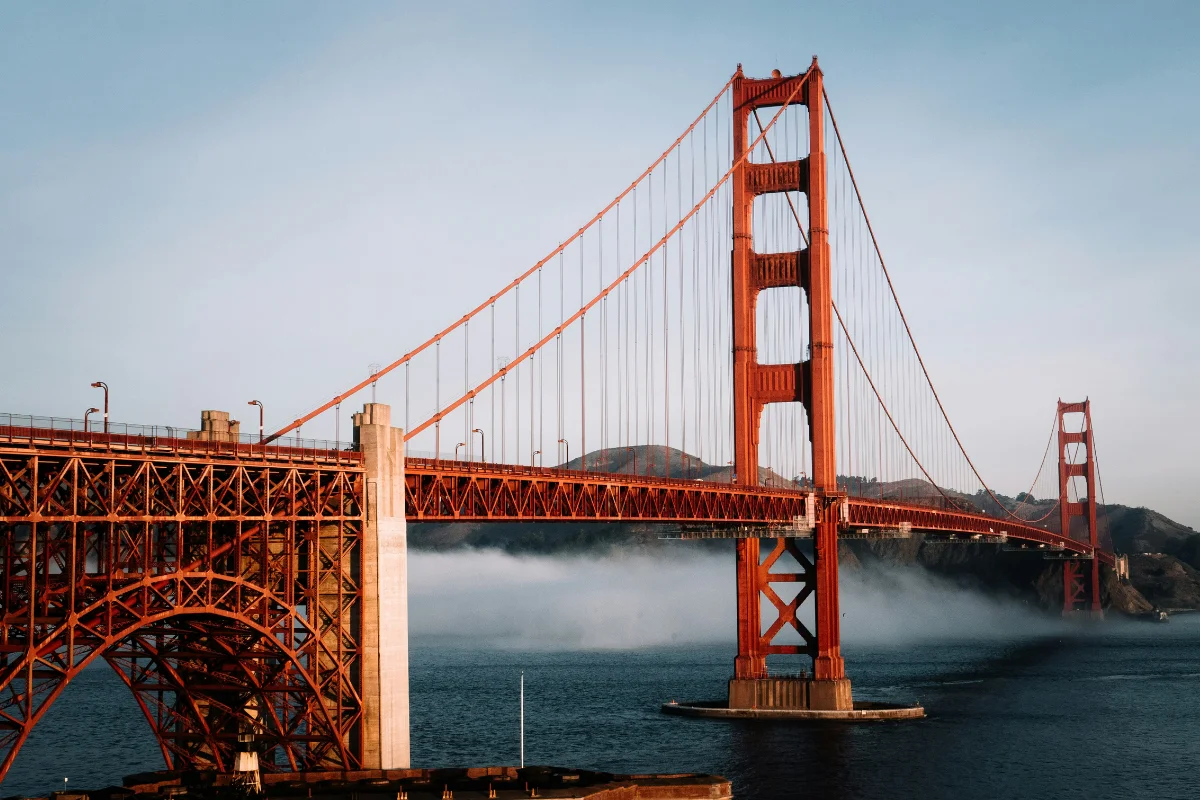 Side view of the iconic orange Golden Gate Bridge spanning over blue bay water with low-lying white fog rolling under the bridge frame.