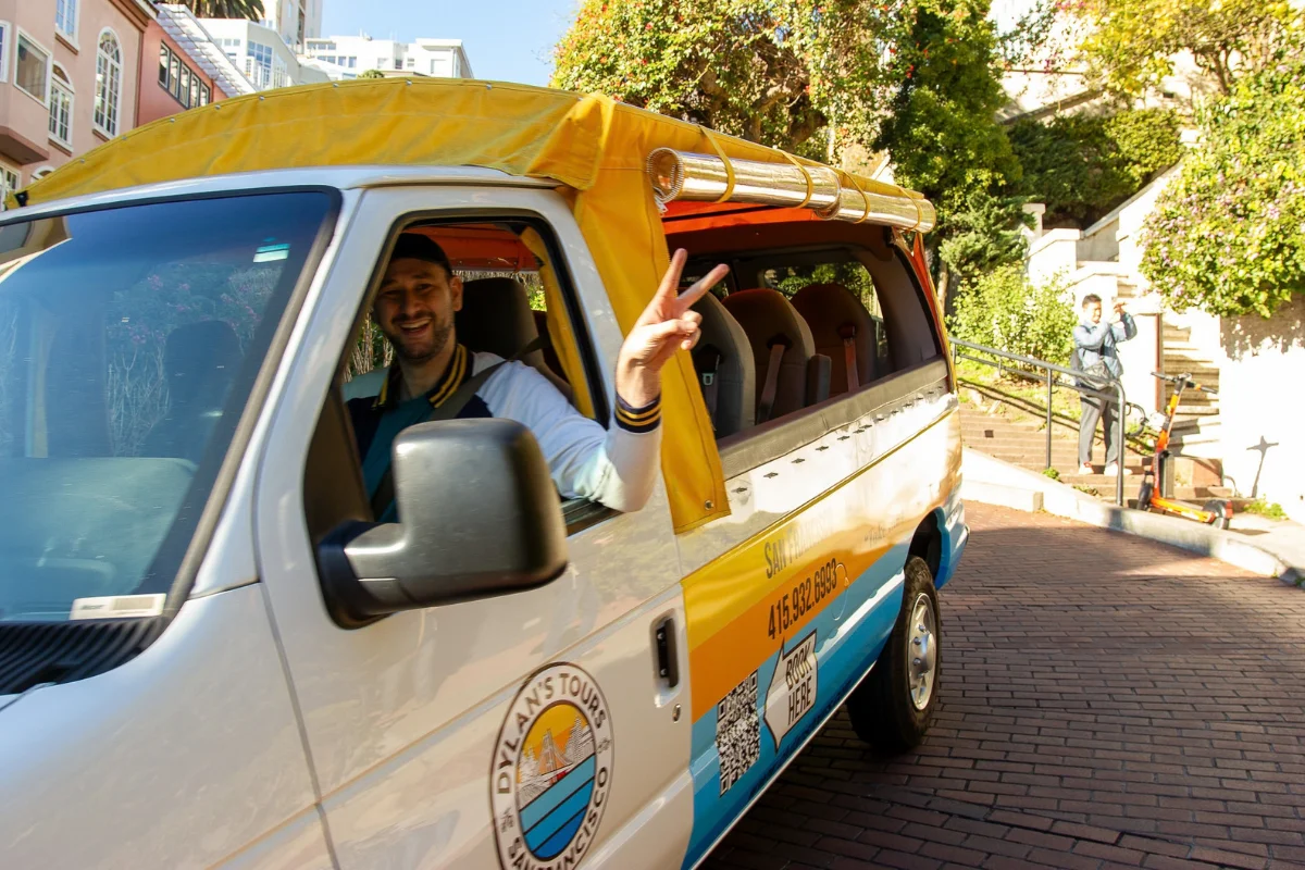 Open-air tour van driver giving a peace sign while driving down a brick street past lush green trees in San Francisco.