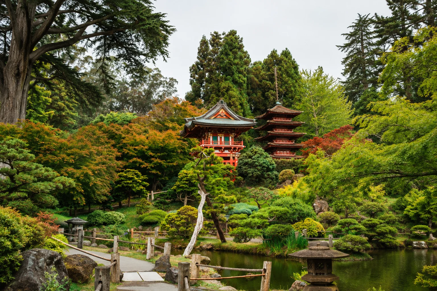 Traditional multi-tiered Japanese pagoda and red temple building surrounded by trees showing green and autumn orange foliage above a calm pond.