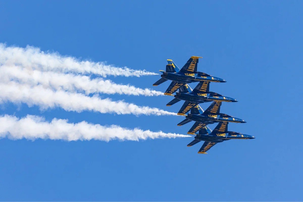 Four blue and yellow US Navy Blue Angels fighter jets flying in tight diagonal formation across a clear blue sky, leaving white smoke trails behind them.