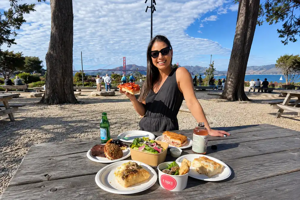 Woman holding a slice of pizza at a picnic table spread with salads, sandwiches, and pastries, with the Golden Gate Bridge behind her